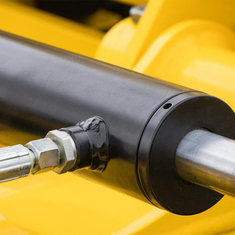 Na zdjęciu widoczny jest siłownik hydrauliczny, czyli element mechaniczny wykorzystywany do generowania siły w jednym kierunku poprzez ruch posuwisto-zwrotny. Korpus siłownika jest czarny, a z jednej strony wysuwa się metalowy tłok. Do urządzenia podłączony jest przewód hydrauliczny, który dostarcza ciecz roboczą.