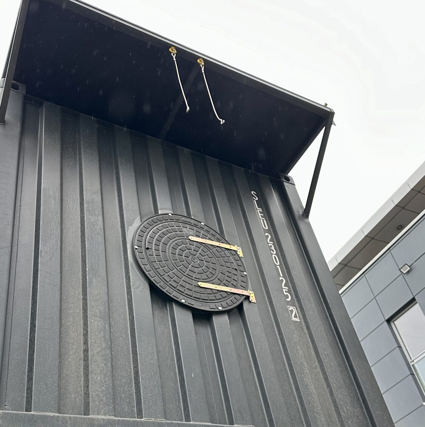 Na zdjęciu widzimy zewnętrzną ścianę kontenera treningowego, prawdopodobnie wykorzystywanego w szkoleniach techników wiatrowych, takich jak kursy GWO. Kontener jest ciemny, stalowy i ma charakterystyczny industrialny wygląd.