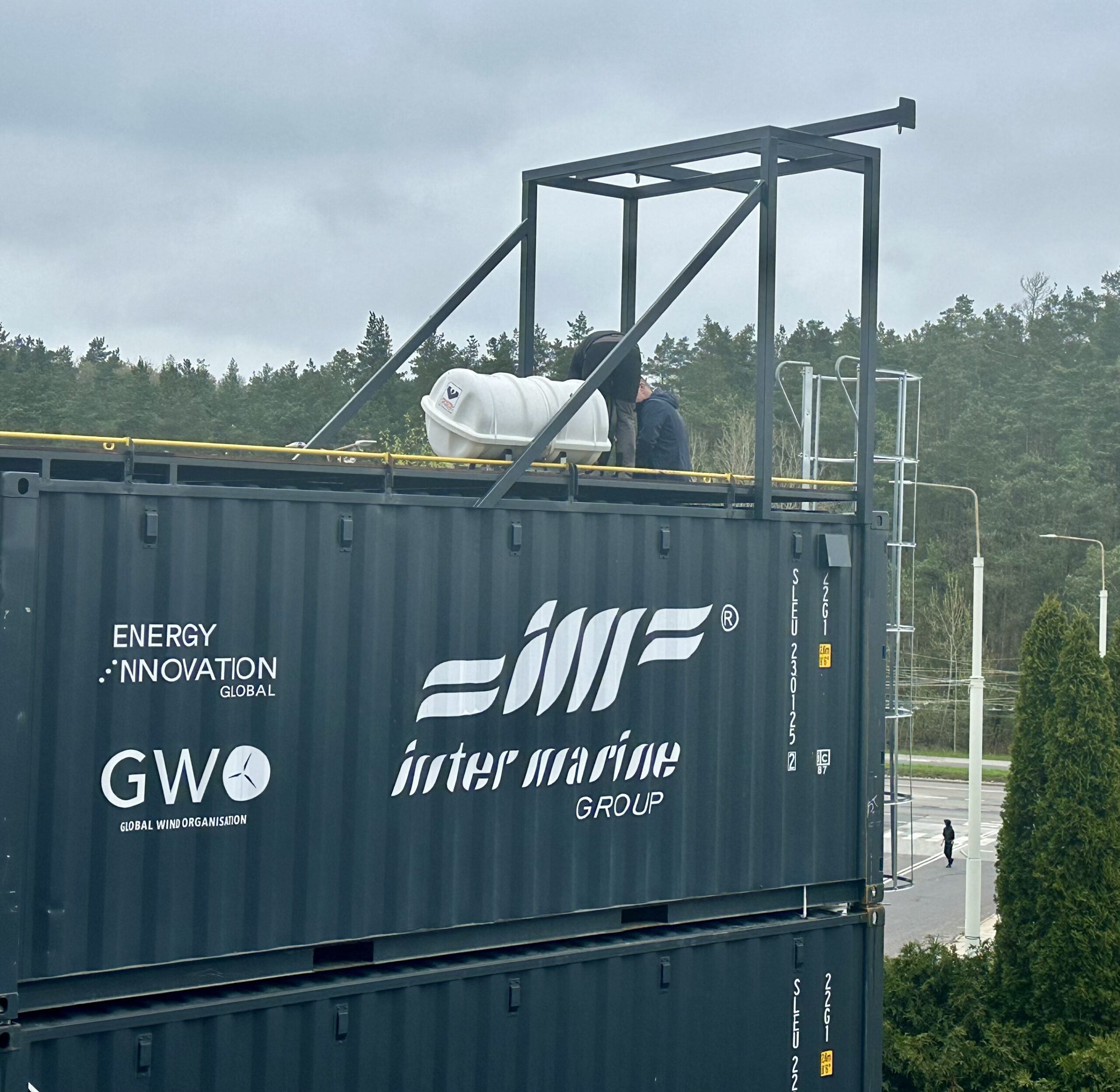 Na zdjęciu widać dwa ustawione jeden na drugim kontenery techniczne z logo Inter Marine Group, Energy Innovation Global oraz GWO (Global Wind Organisation). Na górnym kontenerze znajduje się stalowa konstrukcja, zbiornik oraz elementy wyposażenia technicznego, a z boku zamontowano metalową drabinę prowadzącą na jego górę. W tle widoczny jest zalesiony teren i infrastruktura drogowa.