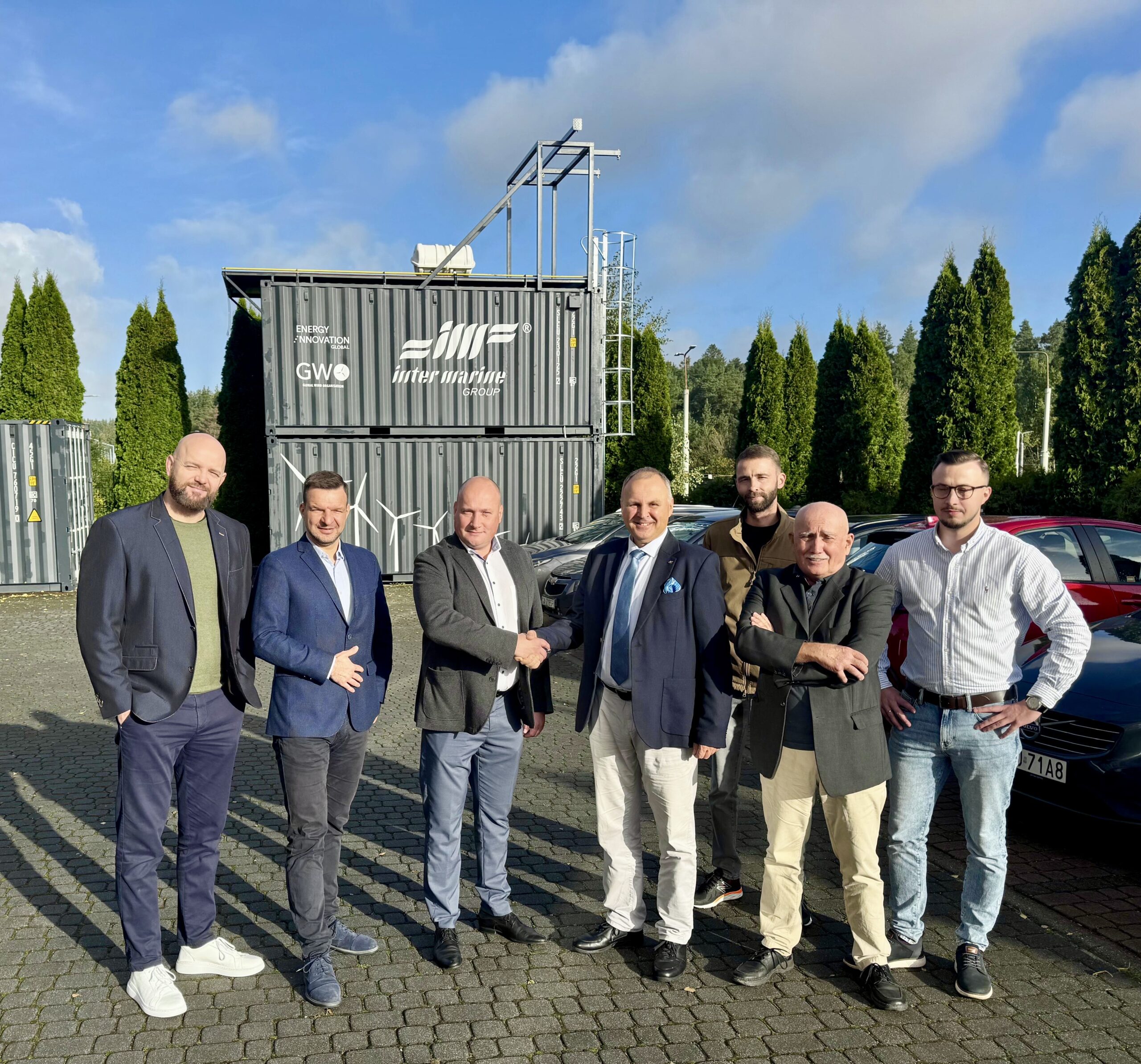 Na zdjęciu znajduje się siedmiu mężczyzn stojących przed kontenerami technicznymi należącymi do Inter Marine Group i oznaczonymi również logotypem GWO (Global Wind Organisation) oraz Energy Innovation. Mężczyźni pozują do wspólnego zdjęcia na zewnątrz, na utwardzonym placu. Ubrani są głównie w marynarki, koszule i spodnie w stylu biznesowo-casualowym. Jeden z nich podaje rękę stojącemu obok mężczyźnie, pozostali patrzą w stronę aparatu.