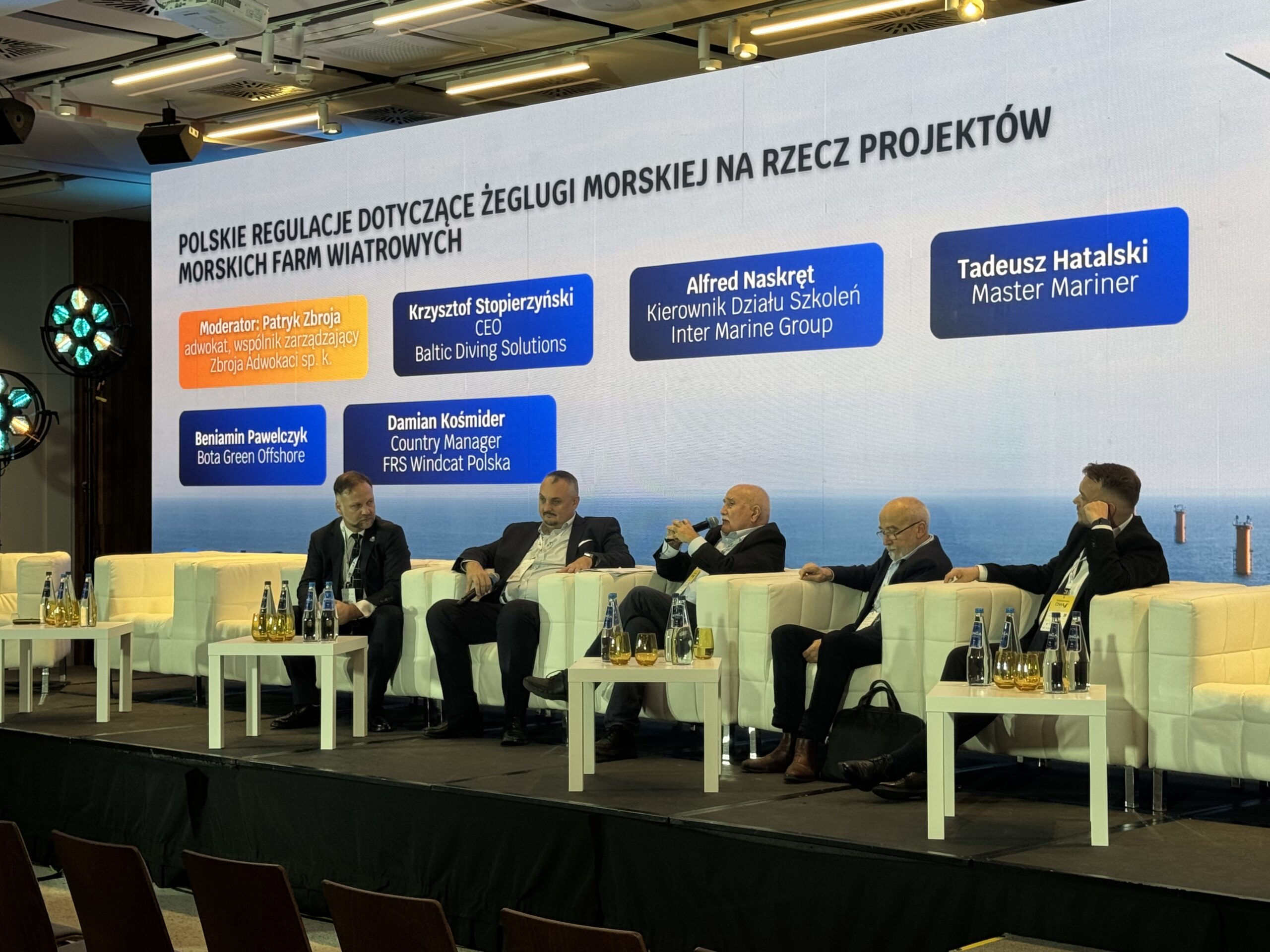 Na zdjęciu widać panel dyskusyjny podczas wydarzenia branżowego poświęconego żegludze morskiej i morskiej energetyce wiatrowej. Na scenie siedzi pięciu uczestników, którzy prowadzą rozmowę przed publicznością. Na dużym ekranie za nimi widnieje temat „Polskie regulacje dotyczące żeglugi morskiej na rzecz projektów morskich farm wiatrowych” oraz informacje o moderatorze i panelistach, reprezentujących m.in. Baltic Diving Solutions, Inter Marine Group, FRS Windcat Polska i Bota Green Offshore.